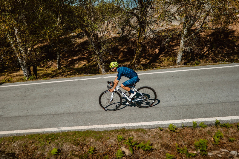 Ciclista con equipación azul marino y casco amarillo fluorescente circulando por una carretera en medio del bosque