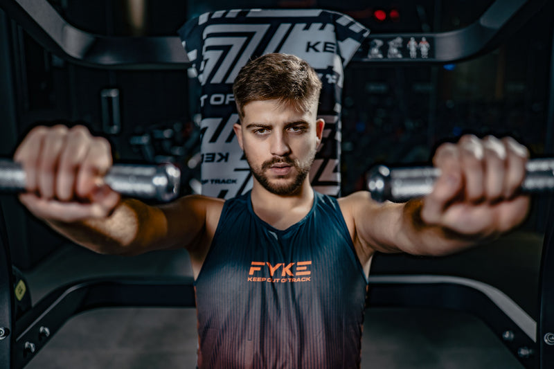 A man in the gym doing chest exercises in a sleeveless Fyke T-shirt.