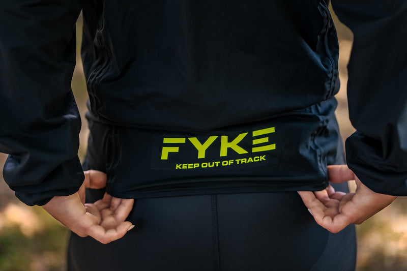 A woman stretches the back of one of Fyke's Running Rain Jackets, showing the brand's logo.